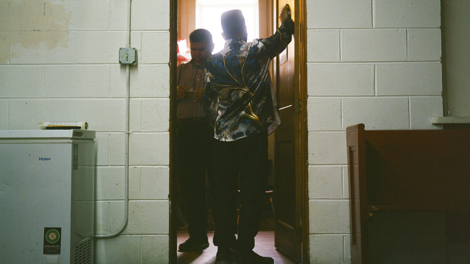 Mohamed Anwar looks at an item in his hand while standing inside an open wood door with another person facing him, with sunlight illuminating them from an open window in the background and a painted concrete-block wall with an appliance and wood pew on either side of the doorway in the foreground.