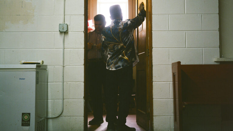 Mohamed Anwar looks at an item in his hand while standing inside an open wood door with another person facing him, with sunlight illuminating them from an open window in the background and a painted concrete-block wall with an appliance and wood pew on either side of the doorway in the foreground.