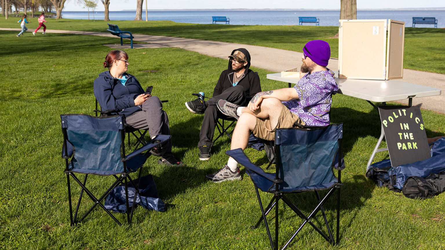 Elaine Koch, Russell Koch and Nikolas DeGroot sit in folding camp chairs arranged in a semi-circle, with two empty folding camp chairs facing them, on a lawn in front of a folding table with a wood box on its surface and a sign reading Politics in the Park leaning against its legs, with two children running next to a sidewalk behind them, and with trees and metal park benches facing a lake in the background. Elaine Koch, Russell Koch and Nikolas DeGroot sit in folding camp chairs arranged in a semi-circle, with two empty folding camp chairs facing them, on a lawn in front of a folding table with a wood box on its surface and a sign reading Politics in the Park leaning against its legs, with two children running next to a sidewalk behind them, and with trees and metal park benches facing a lake in the background.