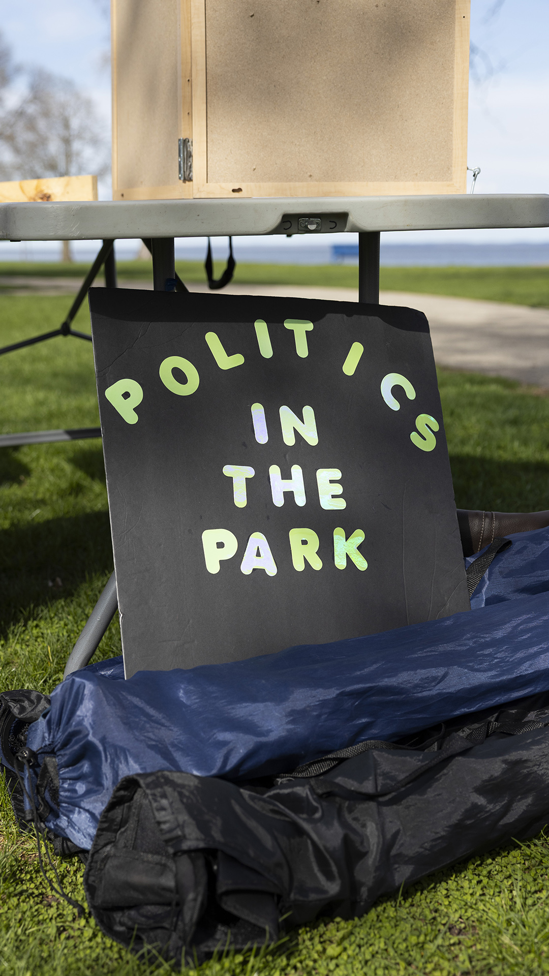 A sign reading "Politics in the Park" is propped against the legs of a folding table and multiple bagged folding camp chairs on a lawn, with an out-of-focus sidewalk, trees and lake in the background.