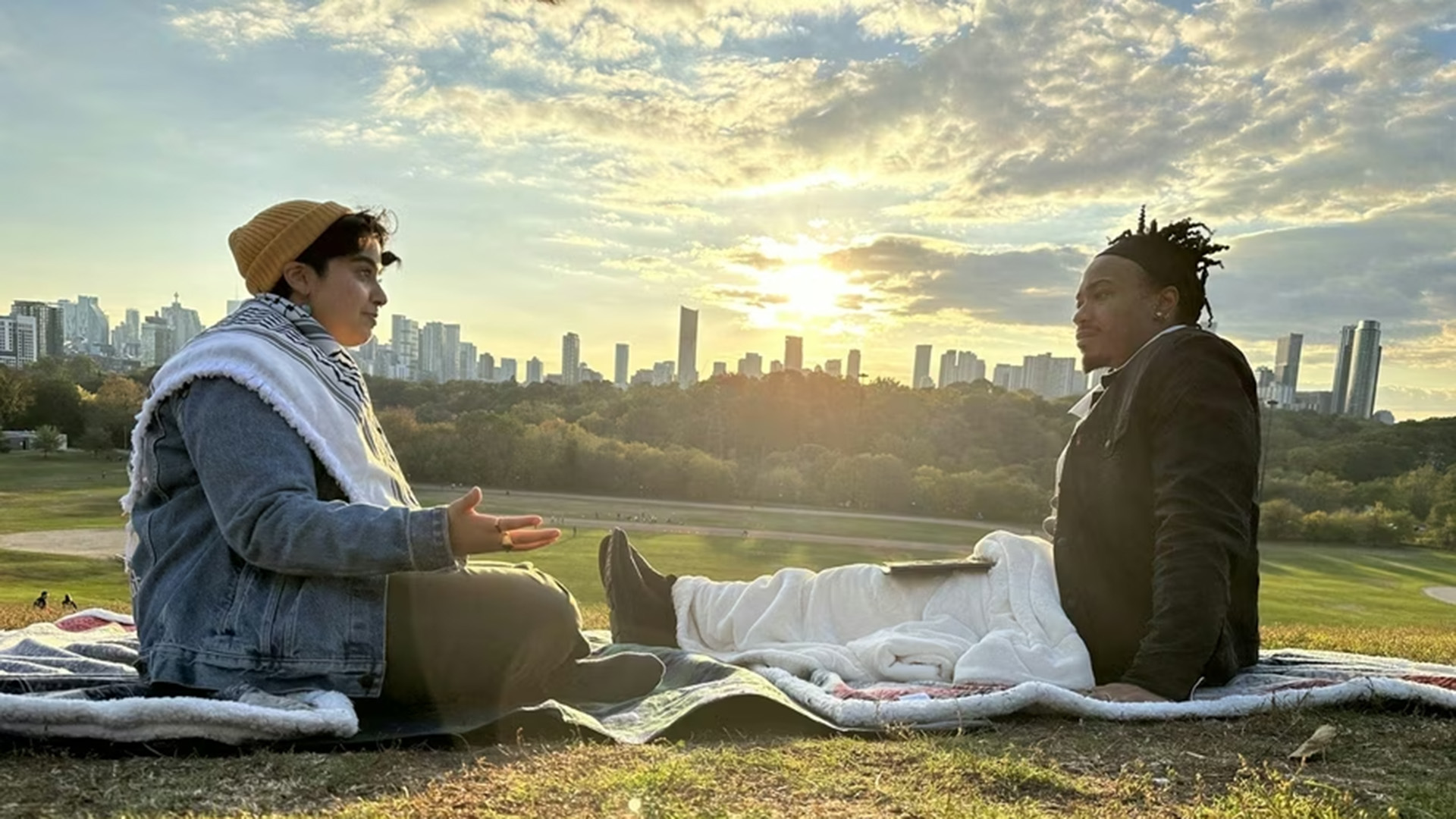 Two people sit on a blanket on a grassy hillside in conversation at sunset, with a city skyline and glowing clouds stretching behind them — a still from PBS Digital Studios' Brave Spaces.