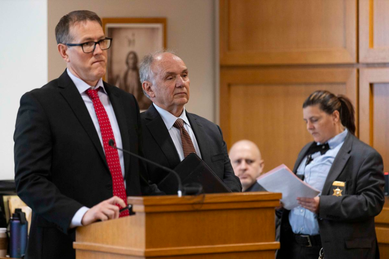 Two men in suits stand at a wooden courtroom lectern with a microphone as a uniformed deputy reads documents beside them.