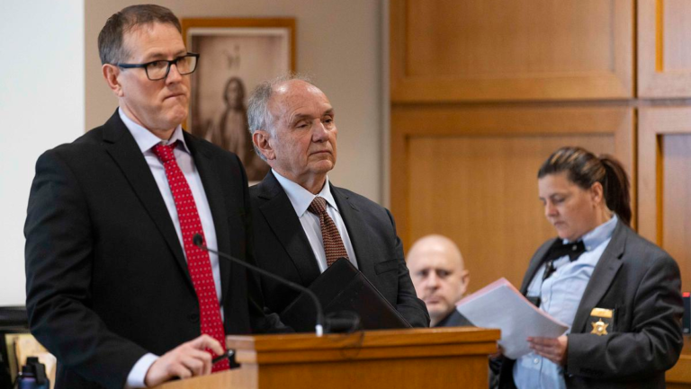 Two men in suits stand at a wooden courtroom lectern with a microphone as a uniformed deputy reads documents beside them.