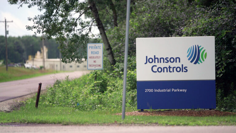 A sign with the words Johnson Controls, a circular logo and an address stands in a lawn next to an intersection in front of a sign reading Private Road and Absolutely No Trespassing, with out-of-focus trees, power lines and buildings in the background.