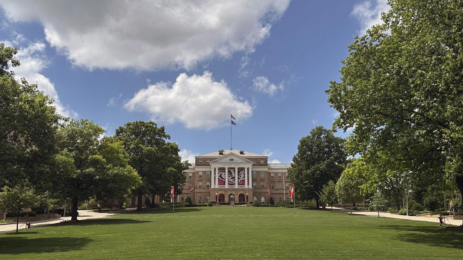 The U.S. and Wisconsin flags fly from a flagpole on top of a multi-story masonry building with a pillar and pediment façade framing a large graphic of the face of the Bucky Badger collegiate mascot, with trees on both sides of a hillside lawn in the foreground under a partly cloudy sky.