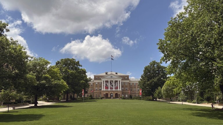The U.S. and Wisconsin flags fly from a flagpole on top of a multi-story masonry building with a pillar and pediment façade framing a large graphic of the face of the Bucky Badger collegiate mascot, with trees on both sides of a hillside lawn in the foreground under a partly cloudy sky.