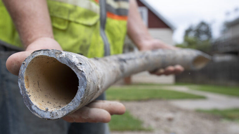 A worker holds a lead service line while standing on a road, with a curb, front yards and houses out-of-focus in the background. A worker holds a lead service line while standing on a road, with a curb, front yards and houses out-of-focus in the background.