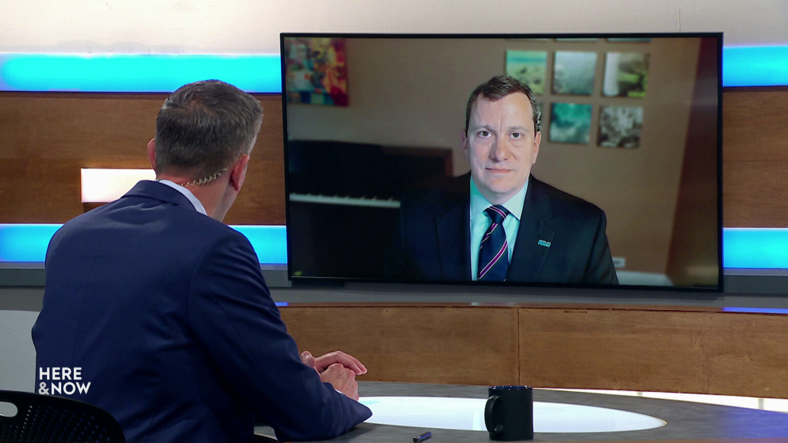 Shawn Johnson sits at a desk on the Here & Now set and faces a video monitor showing an image of Sam Cole.
