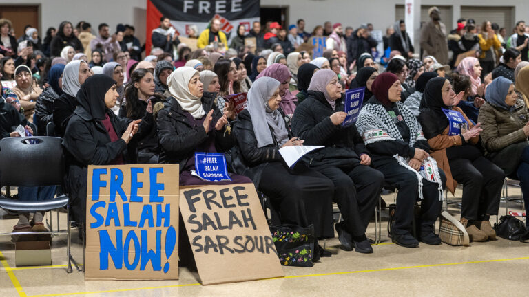 People seated in numerous rows of stacking chairs in a large room hold printed and hand-written signs reading Free Salah Sarsour and Free Salah Now!, with out-of-focus people standing in the background.