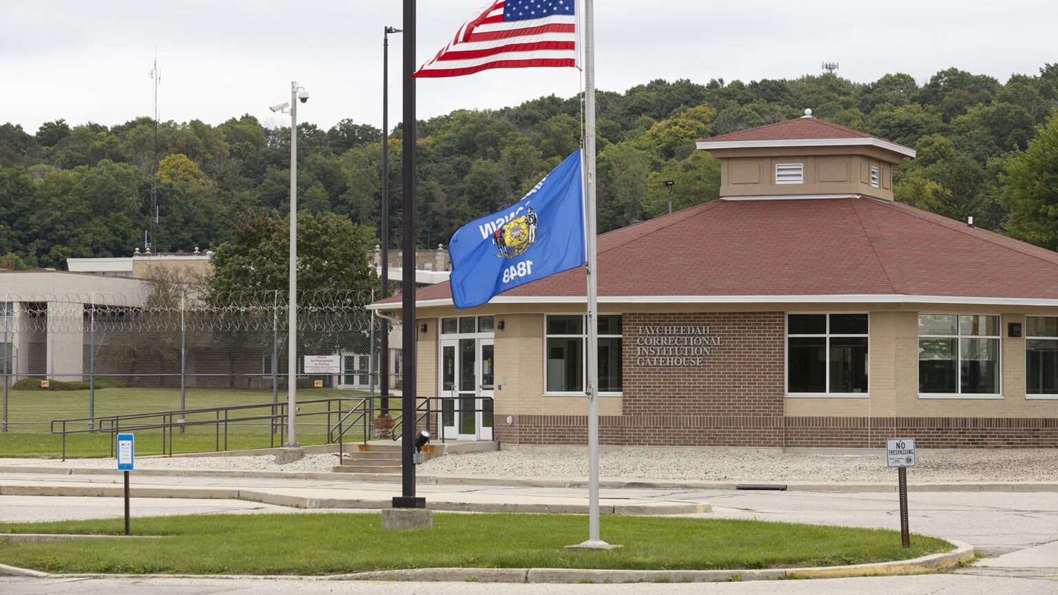 The U.S. and Wisconsin flags fly on a flagpole standing in a lawn island in the middle of a driveway, with a brick building with an octagonal roof and a letter sign reading Taycheedah Correctional Institution Gatehouse next to a chain-link fence topped by razor wire in front of other buildings, with trees on a hill in the background.