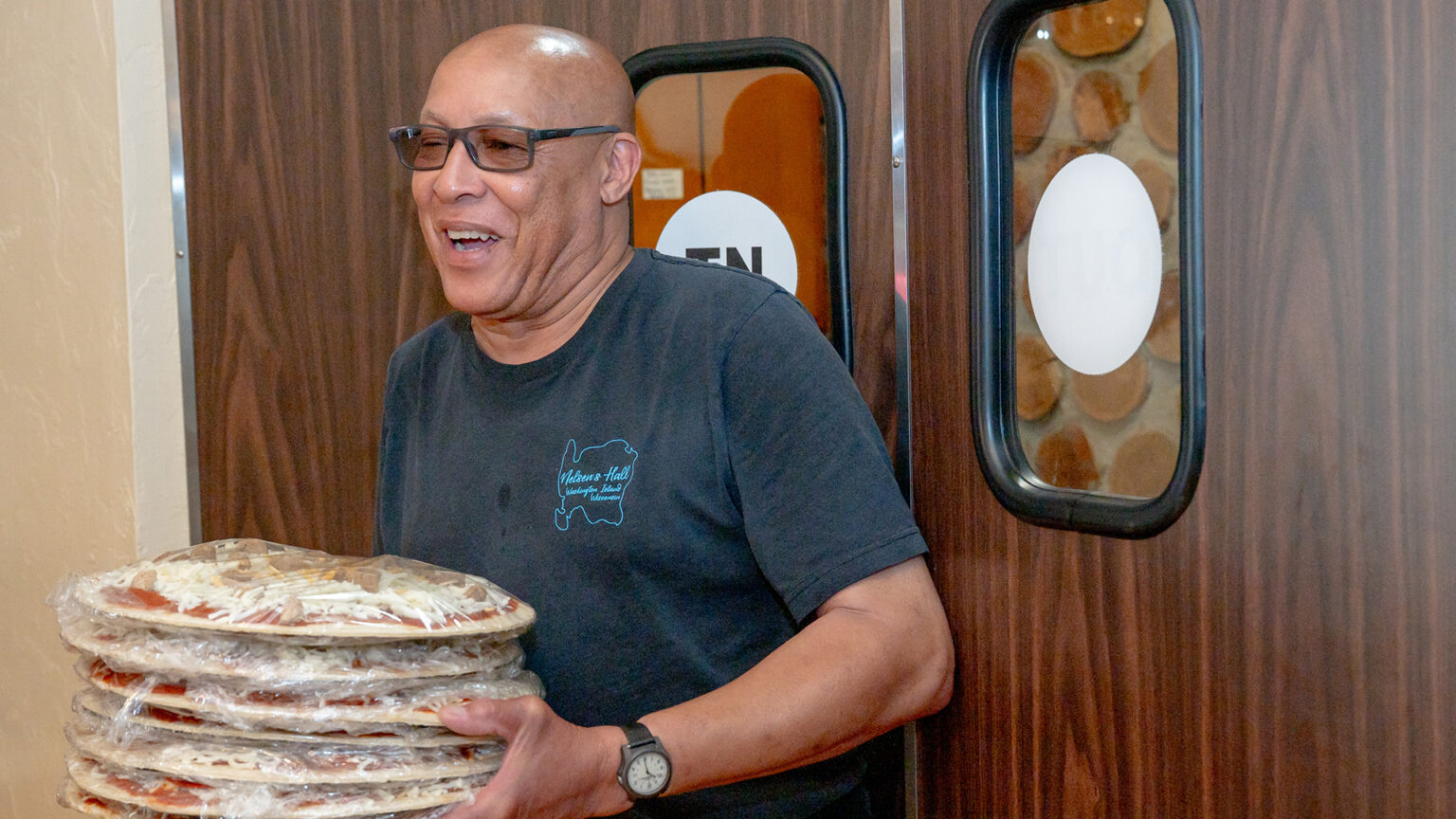 James Carson carries a stack of seven unbaked pizzas wrapped in plastic in front of double doors with a wood laminate finish and windows covered with decals.