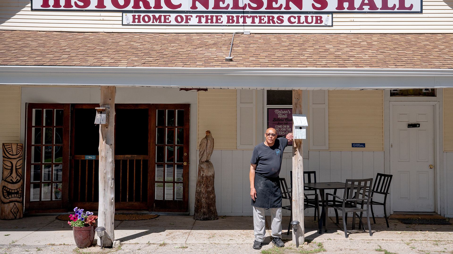 James Cameron poses for a portrait while leaning with one arm against a wood pillar with a wood birdhouse affixed to it under a shingled awning supported by another wood pillar to the side with a flower pot next to its base, with a table with four chairs and multiple open and closed doors and windows behind him and with a sign on the roof reading "Historic Nelsen's Hall" and "Home of the Bitters Club."
