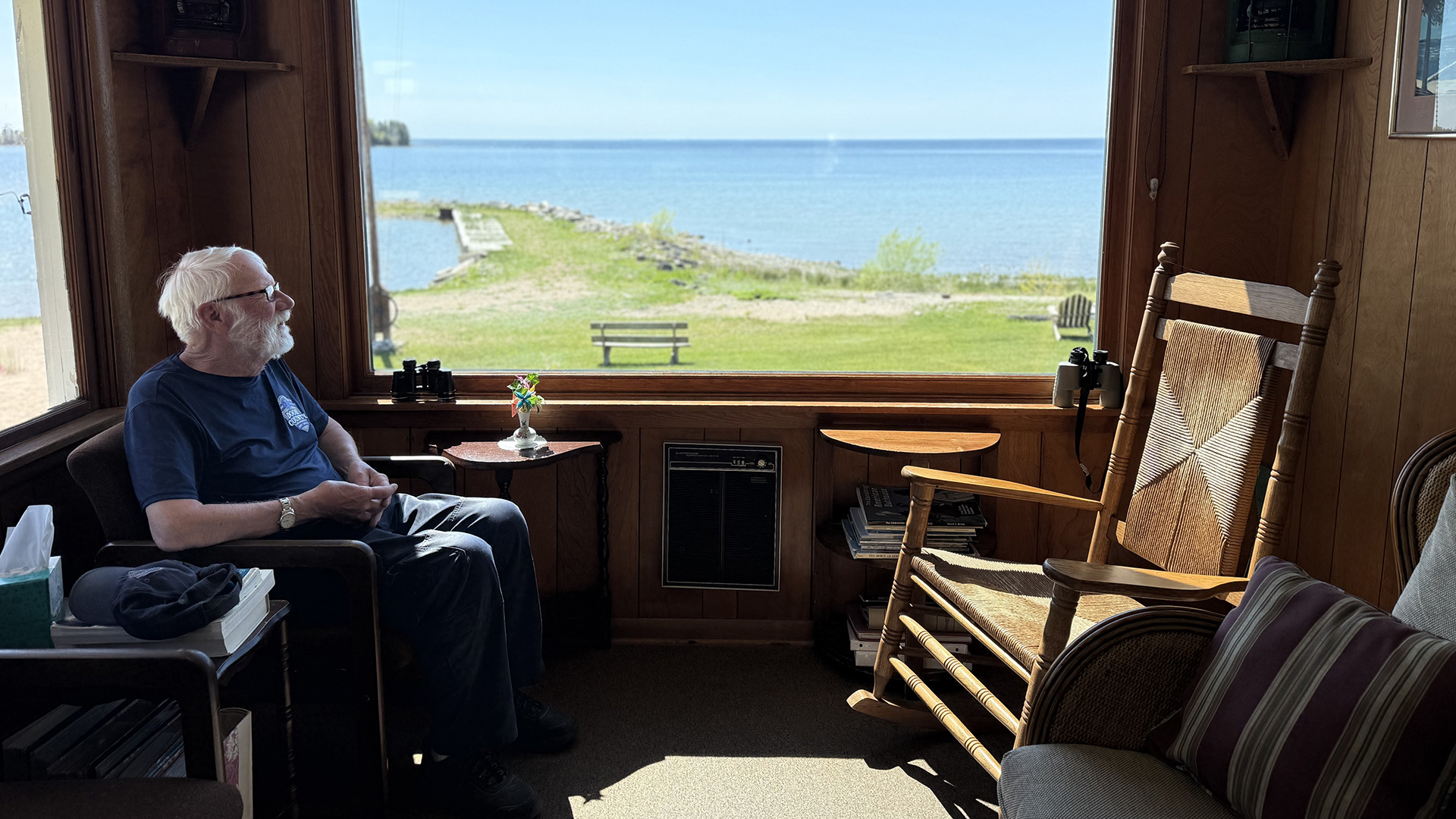 Lee Engstrom sits in an armchair and faces a rocking chair in a room with wood-paneled walls and a large picture window facing a lawn with a bench and chairs and jetty into a lake with waters extending to the horizon.