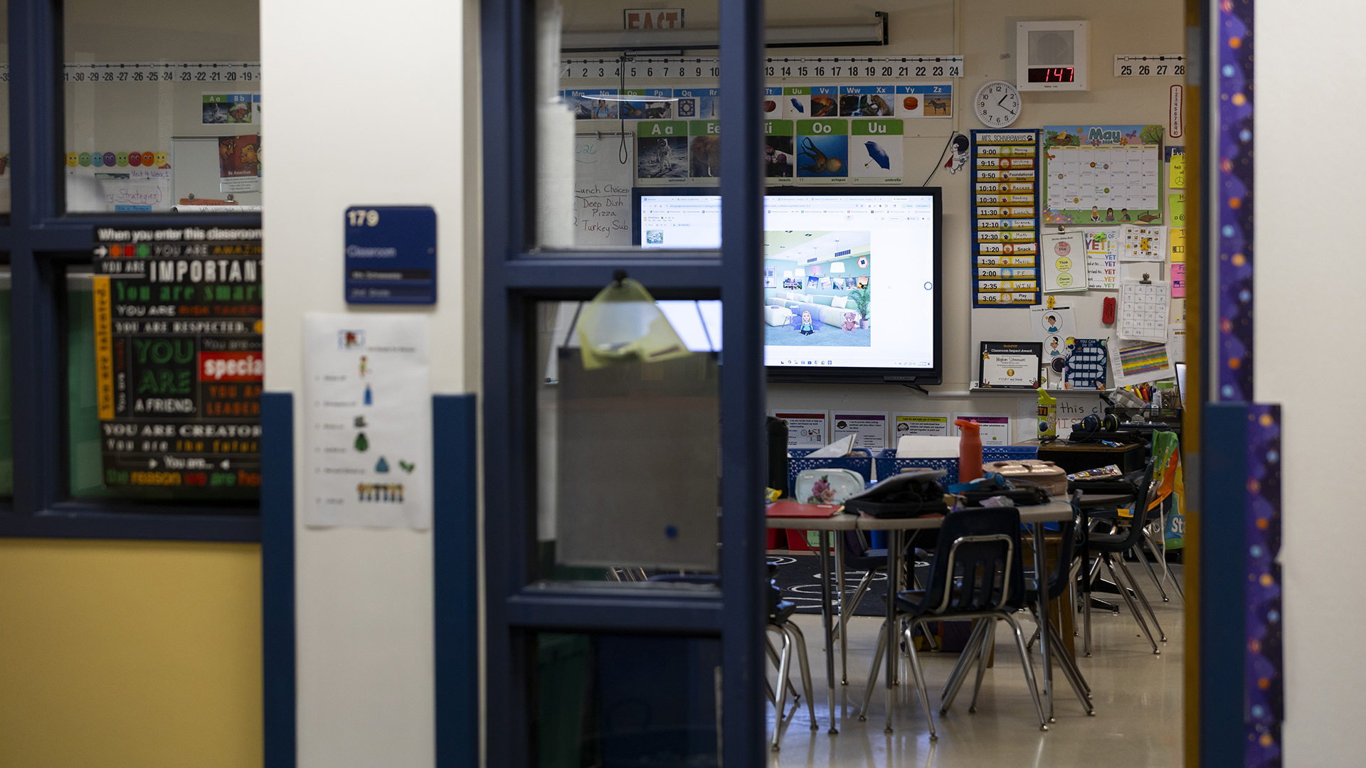 A large wall-mounted monitor is illuminated in a classroom with multiple calendars and teaching tools displayed on its walls and filled multiple low tables surrounded by small chairs, as seen through an open doorway with a glass-paneled doorframe and an out-of-focus room number sign.