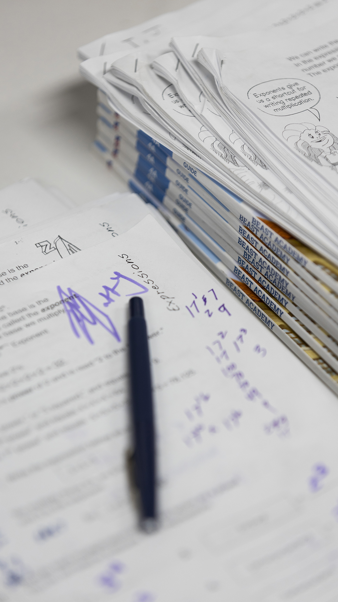 An out-of-focus pen sits on its side on top of a printed worksheet with ink notes written on its surface net to a stack of bound and stapled printed materials.