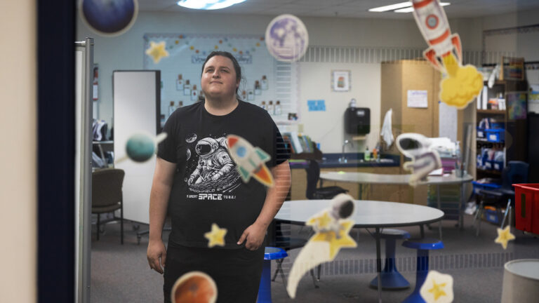Matthew Jacobson stands in a classroom in front of multiple round tables surrounded by stools and chairs, with a room divider, whiteboard, sink and cabinets and shelves along the wall in the background, and with out-of-focus vinyl space-themed decals applied to a glass window in the foreground.