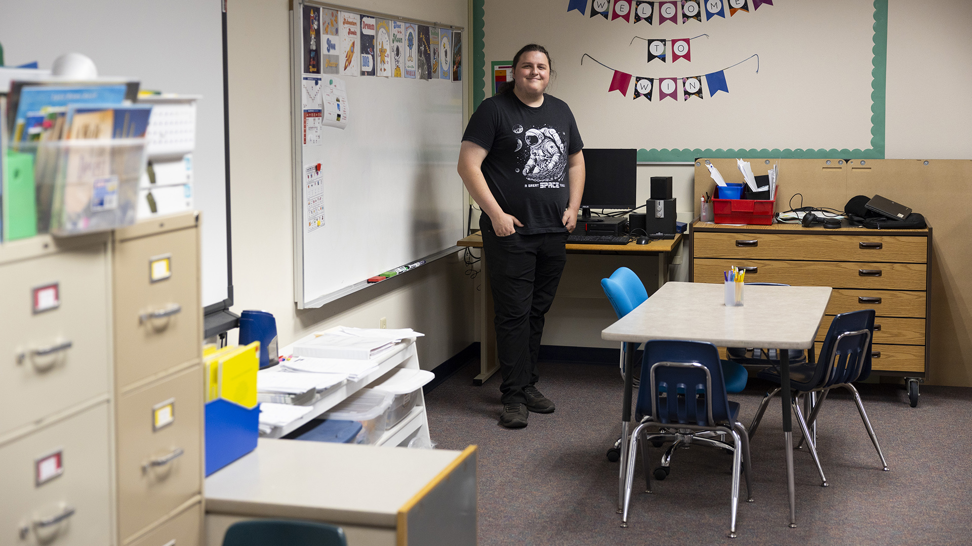 Matthew Jacobson poses for a portrait while standing in the corner of a classroom with a whiteboard, desk with a computer monitor and keyboard on its surface and wood cabinet behind him, and with a short table surrounded by chairs in front of him and out-of-focus metal filing cabinets to one side in the foreground.