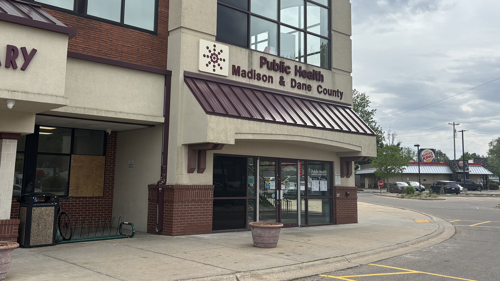 A letter sign reading "Public Health Madison & Dane County" is affixed to the front of a multi-story brick and concrete building above an awning with a metal roof above a set of glass double doors, with a sidewalk with an empty planter in front of the entrance, and with a parking lot, fast food restaurant and trees in the background to one side.