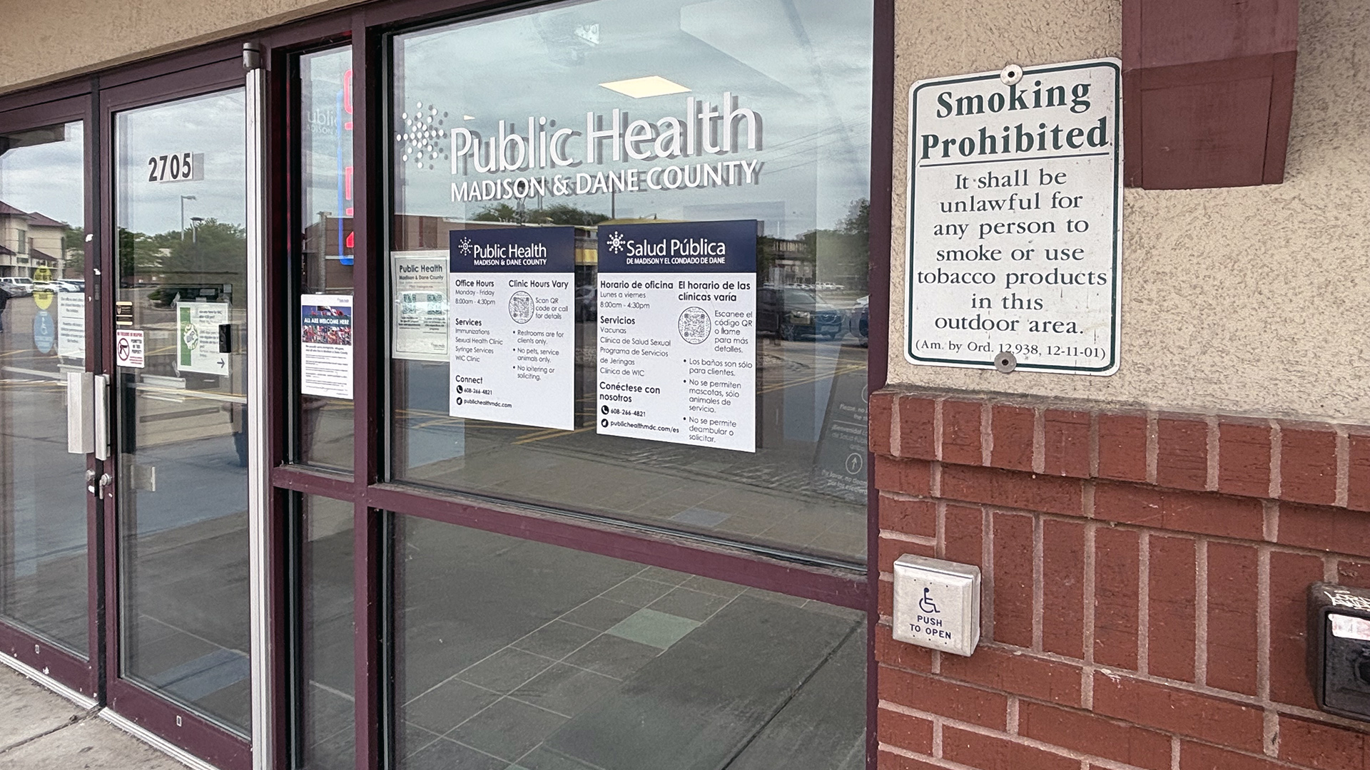 A letter sign reading "Public Health Madison & Dane County" is affixed to the front of a glass window next to a set of glass double doors, with other signs below and to the side, and a metal "Smoking Prohibited" sign affixed to the wall to the side.