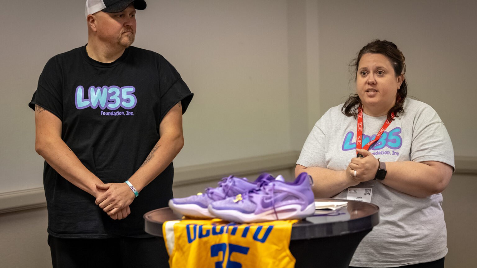 Jamie Weigelt stands and speaks with Jared Weigelt stands next to and looks at her, with a table in between them in the foreground with an out-of-focus pair of shoes and basketball jersey.