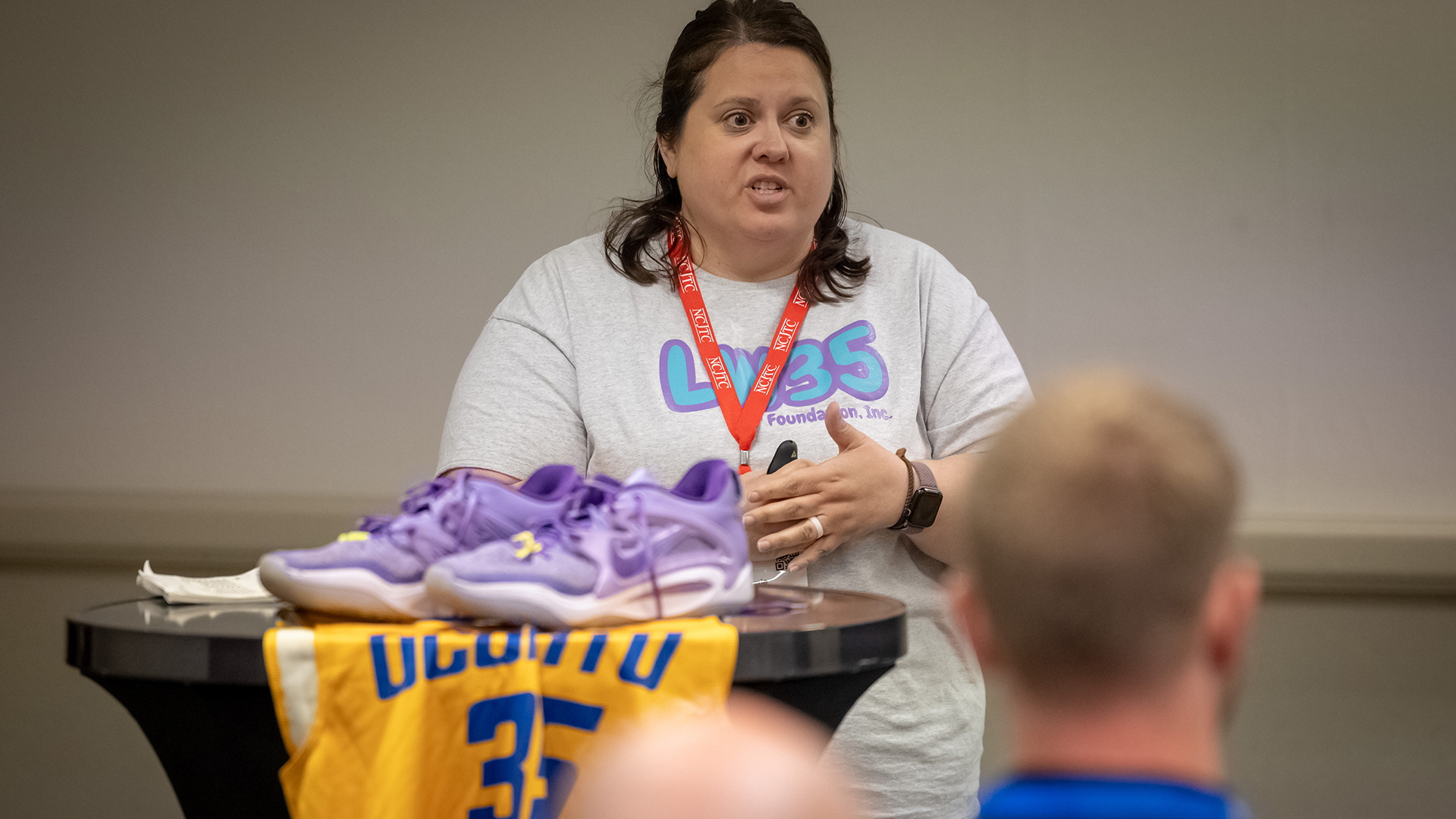 Jamie Weigelt speaks while standing behind a table with an out-of-focus pair of shoes and basketball jersey on its surface, with the out-of-focus backs of two heads in the foreground.