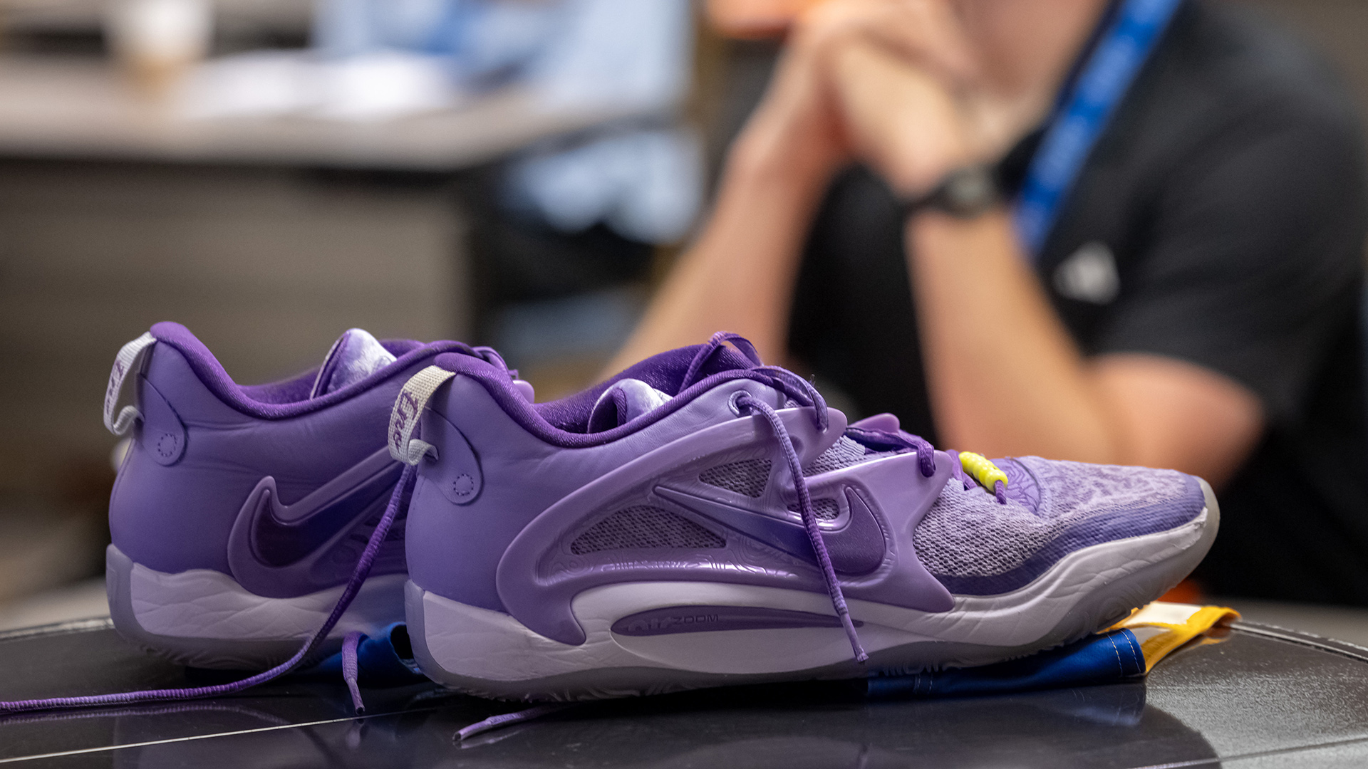 A pair of untied Nike football cleats sit on the surface of a table, with an out-of-focus seated person with steepled arms in the background.