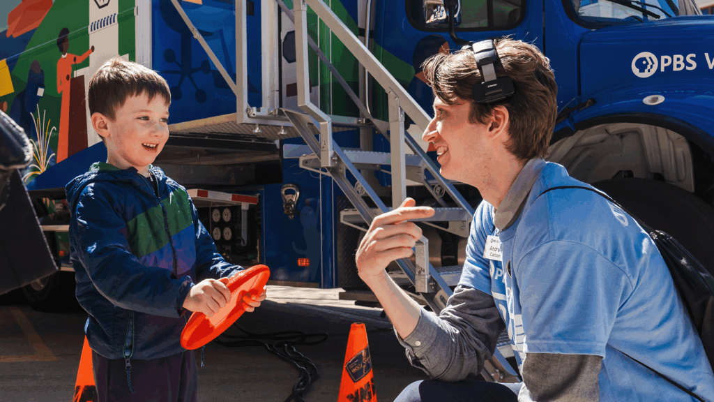A smiling PBS Wisconsin staff member kneels to talk with a young boy holding an orange frisbee in front of the PBS Wisconsin mobile production truck at an outreach event.