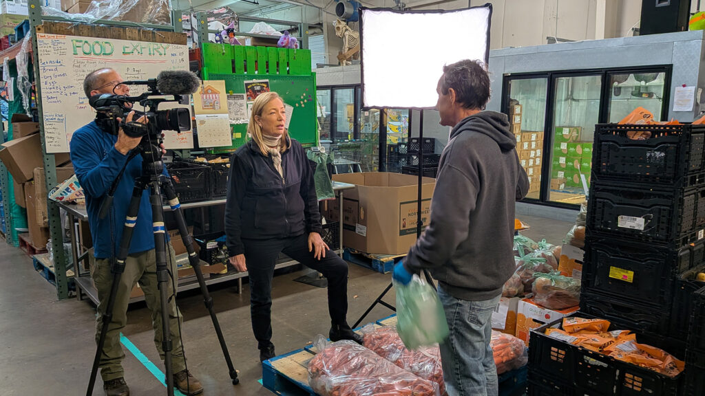 A TV news crew films a reporter interviewing a worker at a food bank warehouse, surrounded by crates of produce and a 