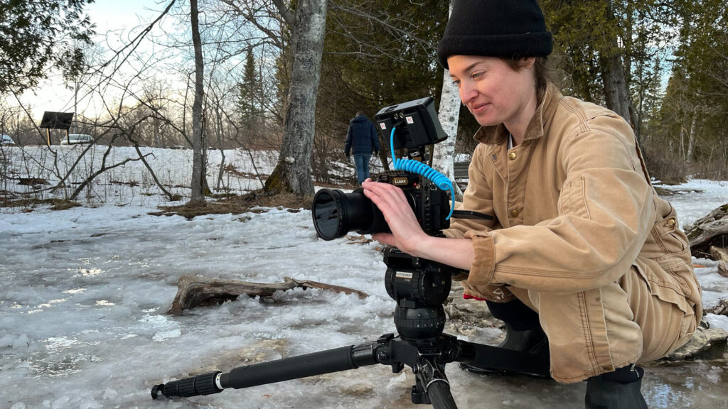 A videographer in a knit cap and canvas jacket adjusts a Lumix camera on a tripod beside a partially frozen creek, filming a wooded winter landscape.