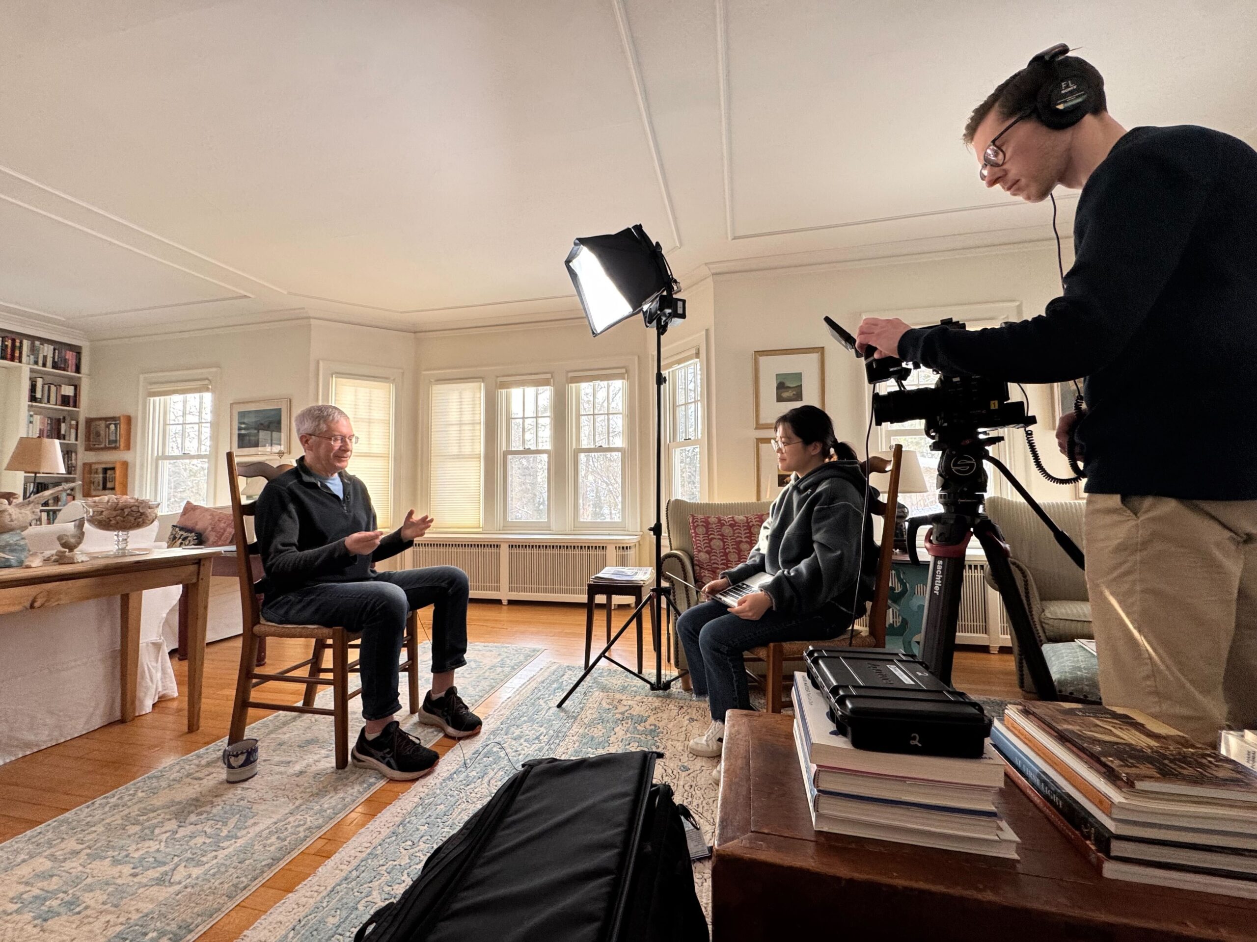 Wisconsin Life student production assistant Heewone Lim interviews a man sitting in a wooden chair in a cozy living room, sitting together with a camera placed on a table in front of them.