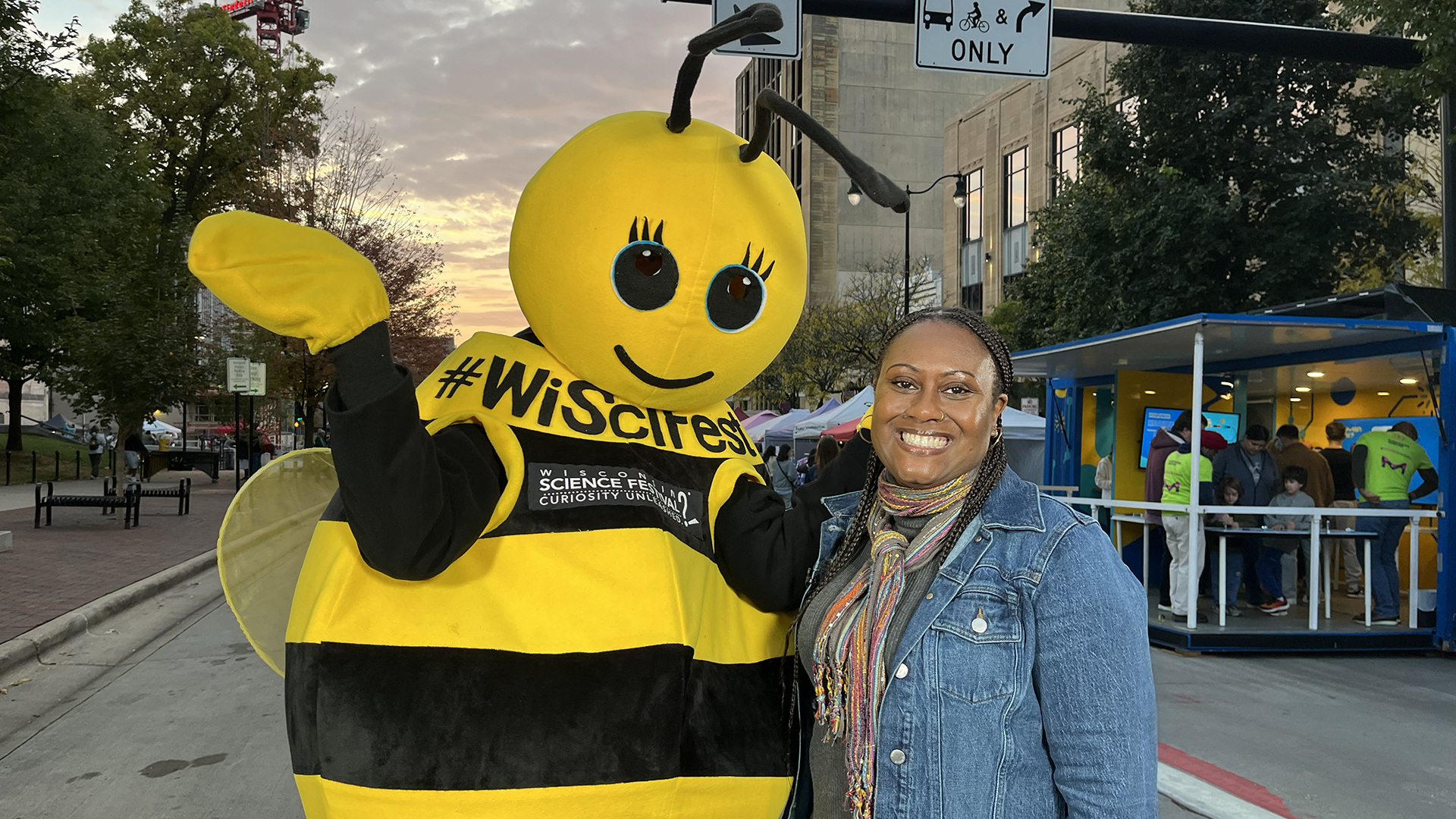 Wisconsin Life host Angela Fitzgerald smiling and standing beside a cheerful bee mascot with "#WiScience" on its outfit. The setting during the 2025 Wisconsin Science Festival is an outdoor urban area at dusk, under a cloudy sky.