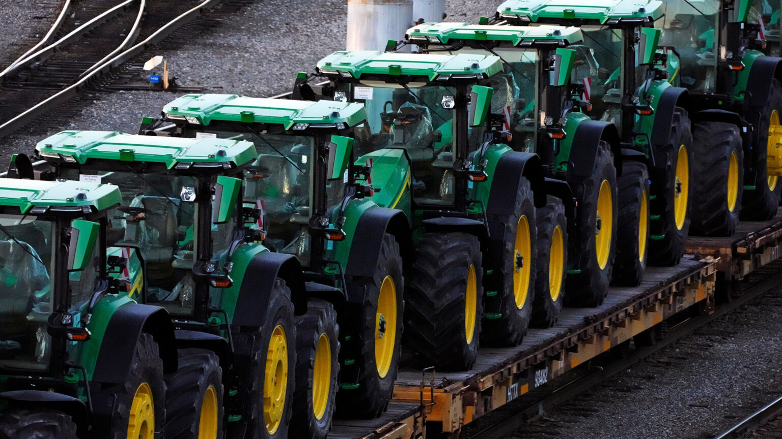 Multiple John Deere tractors are arranged in a line on multiple flat railcars in a railyard with multiple tracks. Multiple John Deere tractors are arranged in a line on multiple flat railcars in a railyard with multiple tracks.