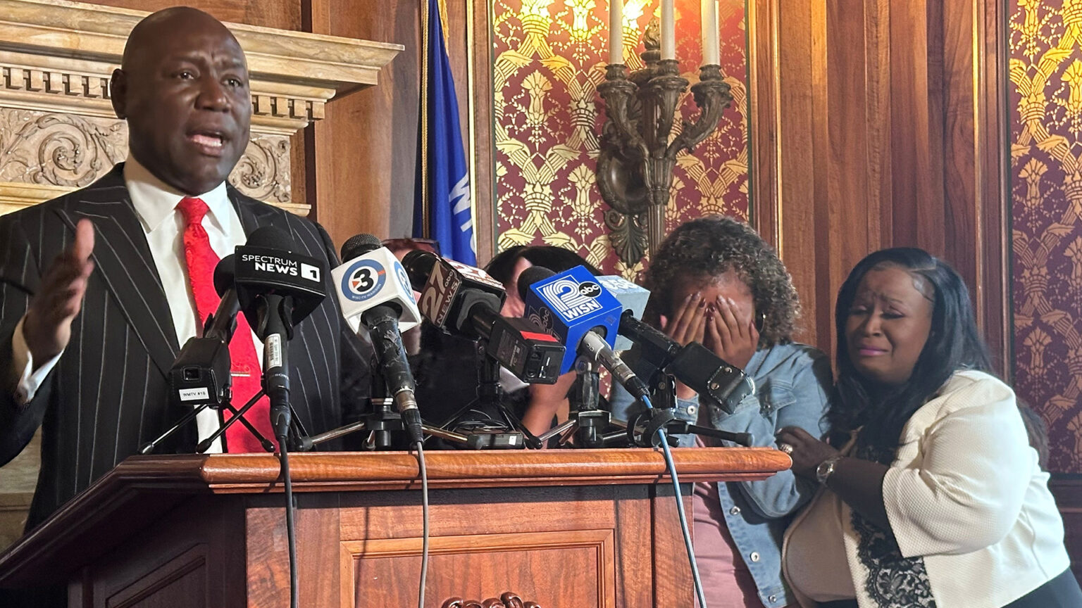 Ben Crump speaks and gestures with his right hand while standing behind a wood podium with multiple microphones with the flags of different media organizations affixed to its surface, with Veronica Ruiz standing to his side holding her face in her hands and Sheila Stubbs holding Ruiz's left arm, in a room with a carved fireplace lintel, the Wisconsin flag, an electric wall sconce and toile wallpaper.
