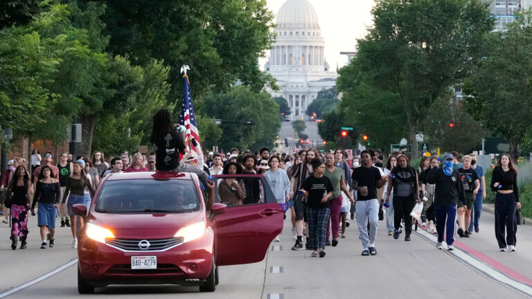A sedan with a driver raising their left arm in a fist out the window and an open driver-side rear door as two people sit on its roof and hold a U.S. flag in the air drives along one side of a boulevard, with a large group of people walking behind across multiple lanes of the street, with trees and multi-story buildings on either side, and the a Beaux-Arts style building with a dome at the top of a low hill in the background. A sedan with a driver raising their left arm in a fist out the window and an open driver-side rear door as two people sit on its roof and hold a U.S. flag in the air drives along one side of a boulevard, with a large group of people walking behind across multiple lanes of the street, with trees and multi-story buildings on either side, and the a Beaux-Arts style building with a dome at the top of a low hill in the background.