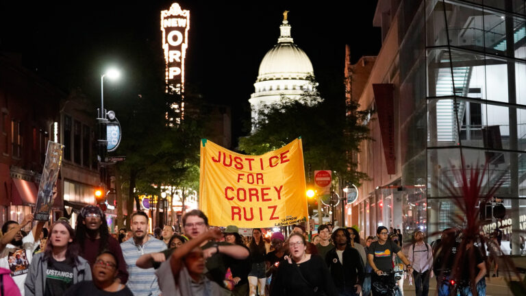 A group of protestors march along a street at night while holding a fabric banner on two poles with the words Justice for Corey Ruiz, with buildings and trees on either side, and with an illuminated theater marquee sign reading New Orpheum and an illuminated dome of Beaux-Arts style building topped by a gilt statue in the background.