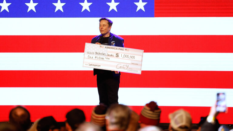 Elon Musk hold an oversized paper check with the words America PAC, pay to the order of Nicholas Jacobs and $1,000,000, with an illuminated U.S. flag on a screen in the background and out-of-focus heads of people facing him in the foreground.
