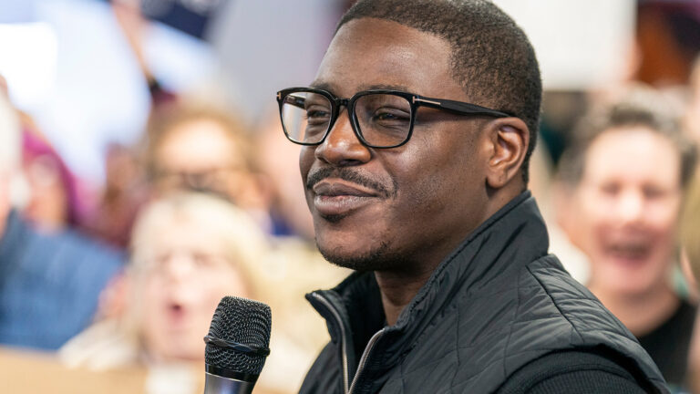 David Crowley smiles while holding a microphone in front of him, with out-of-focus people cheering in the background. David Crowley smiles while holding a microphone in front of him, with out-of-focus people cheering in the background.
