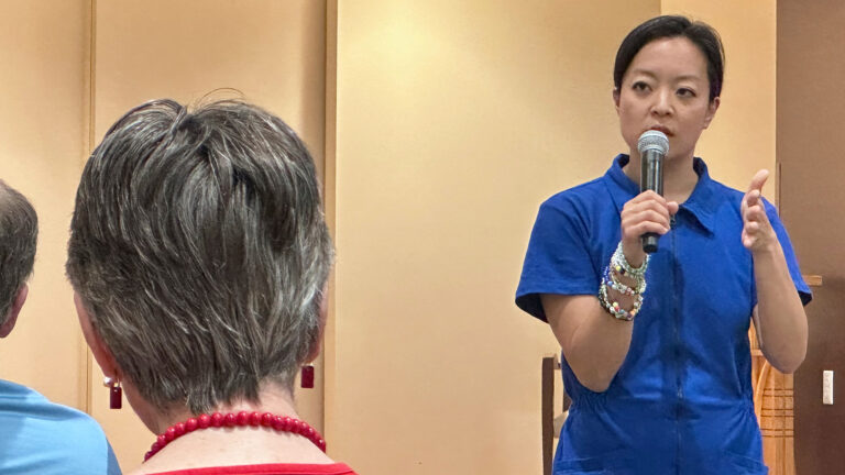 Francesca Hong holds a wireless microphone in her right hand and gestures with her left hand while standing and speaking in a room, with the backs of the heads of seated people facing her in the foreground. Francesca Hong holds a wireless microphone in her right hand and gestures with her left hand while standing and speaking in a room, with the backs of the heads of seated people facing her in the foreground.