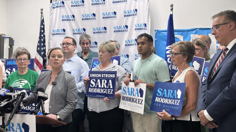 Sara Rodriguez speaks while standing behind a wood podium with multiple microphones with the flags of different media organizations affixed to its top, with multiple people standing behind her holding printed campaign signs reading Sara Rodriguez for Governor and Union Strong Sara Rodriguez, in a room with the U.S. and Wisconsin flags on stands in the background and a backdrop banner showing her campaign logo.