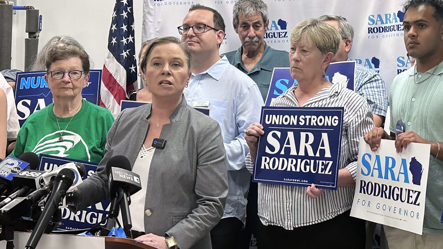 Sara Rodriguez speaks while standing behind a wood podium with multiple microphones with the flags of different media organizations affixed to its top, with multiple people standing behind her holding printed campaign signs reading Sara Rodriguez for Governor and Union Strong Sara Rodriguez, in a room with the U.S. flag in the background and a backdrop banner showing her campaign logo.