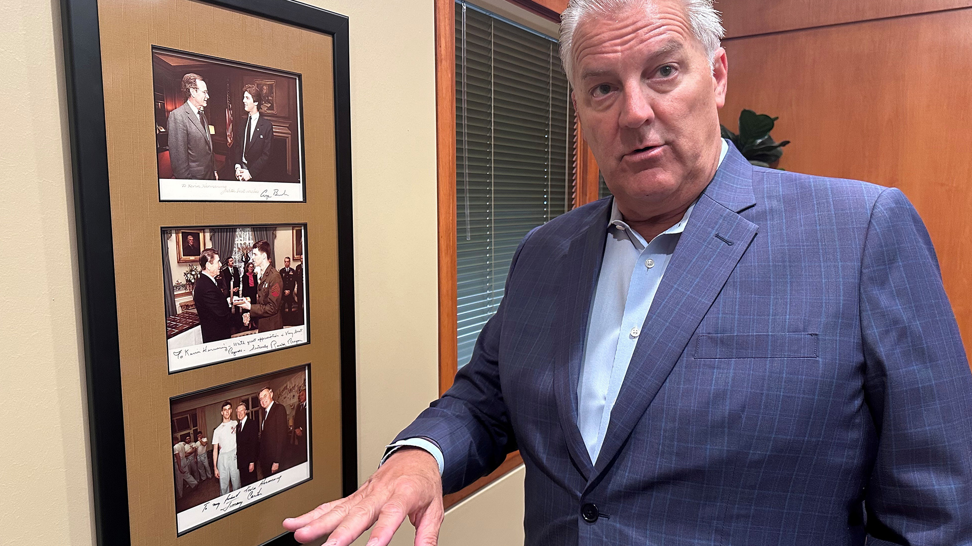 Kevin Hermening points with his right hand toward a frame with three photographs mounted on a wall in a room with a wood door and window with closed blinds in the background.