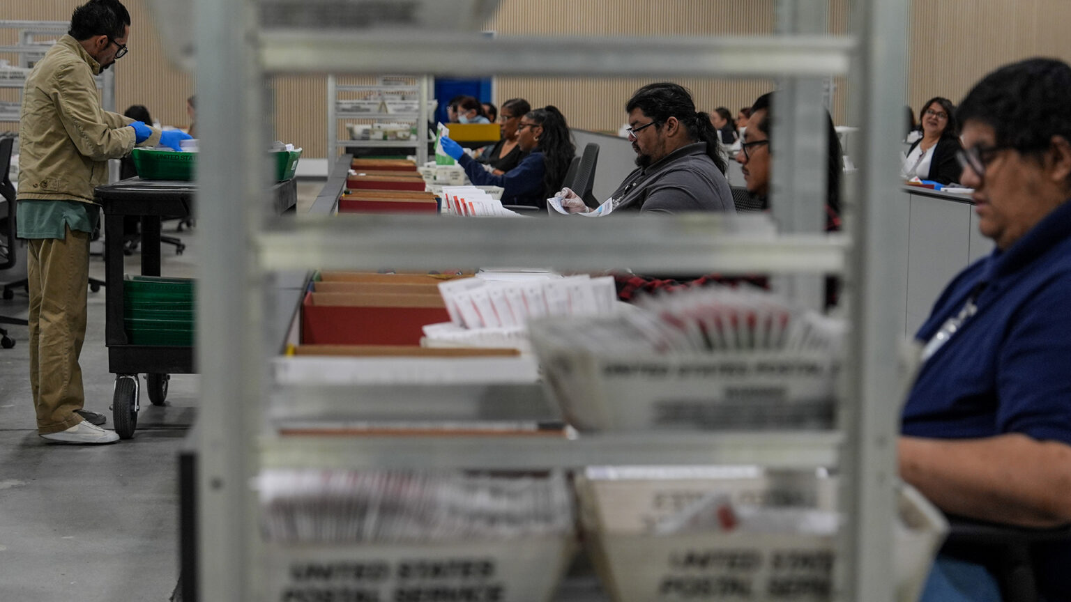 Multiple seated and standing people sort ballot envelopes at a row of tables and next to a rolling cart, with an out-of-focus metal cart with United States Postal Service boxes filled with ballot envelopes stacked on its shelves in the foreground. Multiple seated and standing people sort ballot envelopes at a row of tables and next to a rolling cart, with an out-of-focus metal cart with United States Postal Service boxes filled with ballot envelopes stacked on its shelves in the foreground.