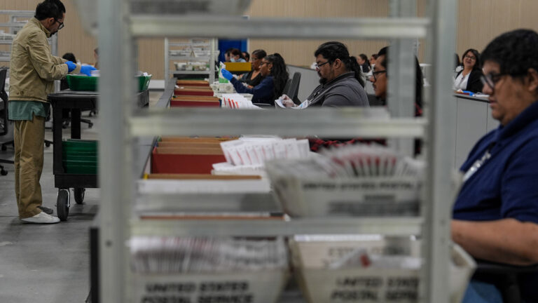 Multiple seated and standing people sort ballot envelopes at a row of tables and next to a rolling cart, with an out-of-focus metal cart with United States Postal Service boxes filled with ballot envelopes stacked on its shelves in the foreground. Multiple seated and standing people sort ballot envelopes at a row of tables and next to a rolling cart, with an out-of-focus metal cart with United States Postal Service boxes filled with ballot envelopes stacked on its shelves in the foreground.