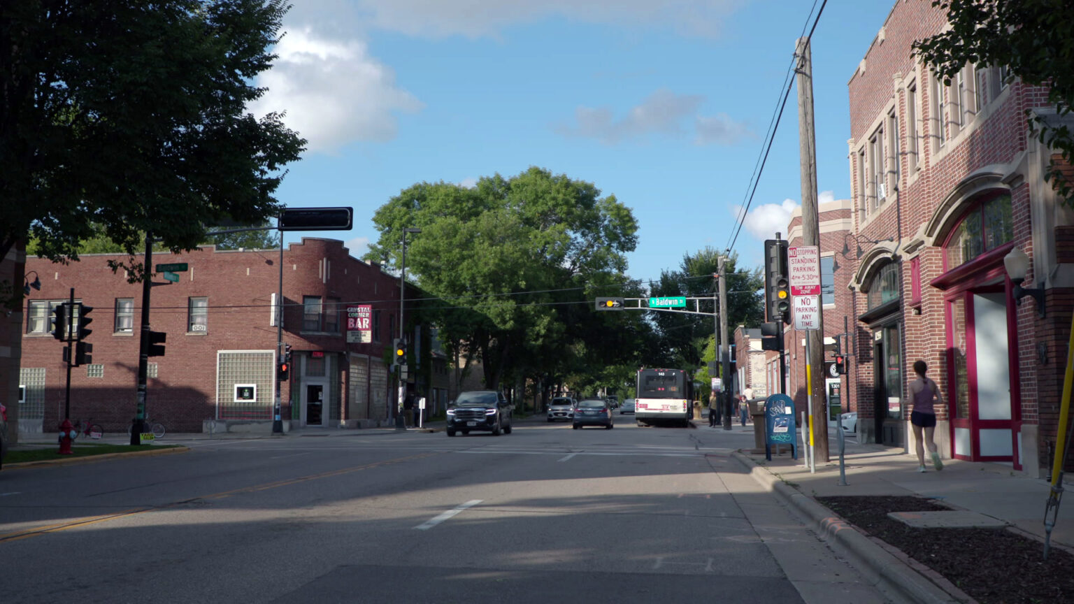 A bus and other vehicles drive along a city street through and past an intersection where traffic lights are illuminated yellow, with brick buildings on either side of the street, with trees and utility poles and traffic signs along the sidewalks. A bus and other vehicles drive along a city street through and past an intersection where traffic lights are illuminated yellow, with brick buildings on either side of the street, with trees and utility poles and traffic signs along the sidewalks.