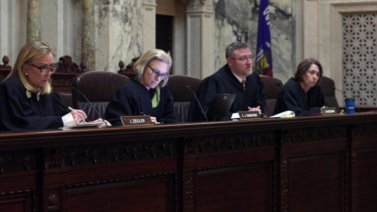 From left to right, Annette Ziegler, Jill Karofsky, Brian Hagedorn and Susan Crawford sit in high-backed leather chairs at a judicial dais, with another row of high-backed wood and leather chairs behind them, in a room with marble masonry and a Wisconsin flag.