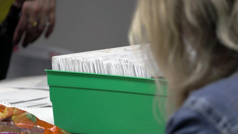 Rows of envelopes sit in.a plastic box, with the out-of-focus hair and shoulders of a person to the side in the foreground, and the out-of-focus hand of a another person to the other side in the background. Rows of envelopes sit in.a plastic box, with the out-of-focus hair and shoulders of a person to the side in the foreground, and the out-of-focus hand of a another person to the other side in the background.