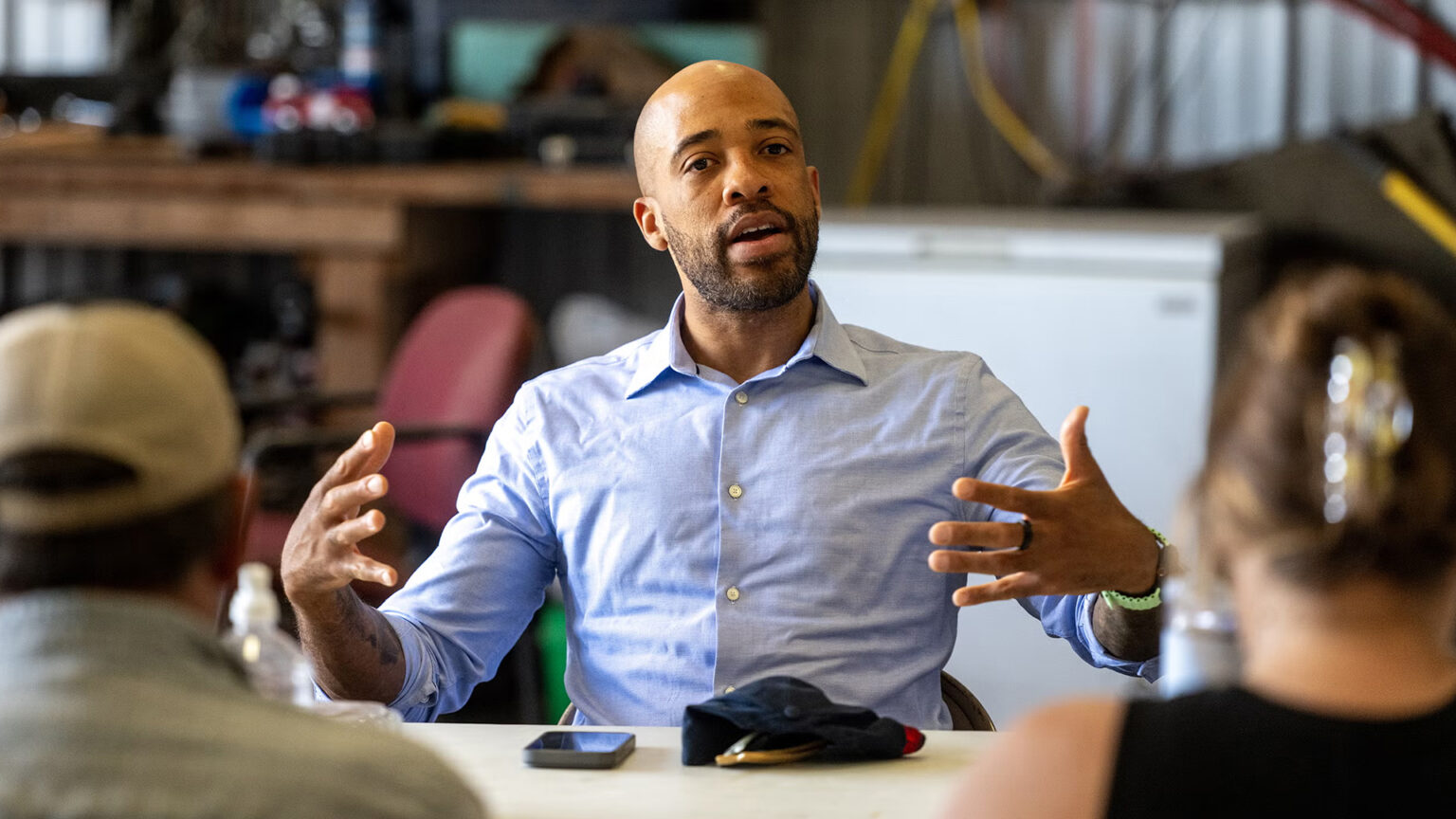 Mandela Barnes gestures with both hands while seated at a table with a phone, hat and sunglasses on its surface in front of him, in a room with an out-of-focus chest freezer and workbench in the background, and with the back of the heads of two seated people who are facing him in the foreground. Mandela Barnes gestures with both hands while seated at a table with a phone, hat and sunglasses on its surface in front of him, in a room with an out-of-focus chest freezer and workbench in the background, and with the back of the heads of two seated people who are facing him in the foreground.