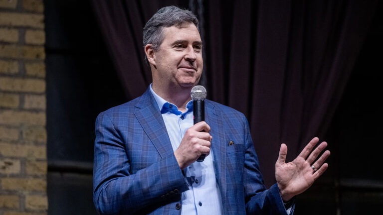 Joel Brennan gestures with his left hand and holds a microphone in his right hand while standing in a room with a brick wall and stage curtain in the background.