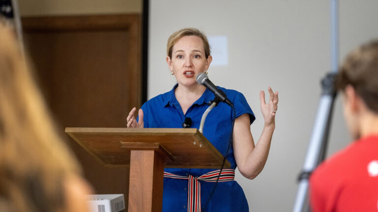 Kelda Roys gestures with both hands while speaking into a microphone mounted to the top of a lectern in a room with a wood door in the background and the out-of-focus heads of people facing her in the foreground.