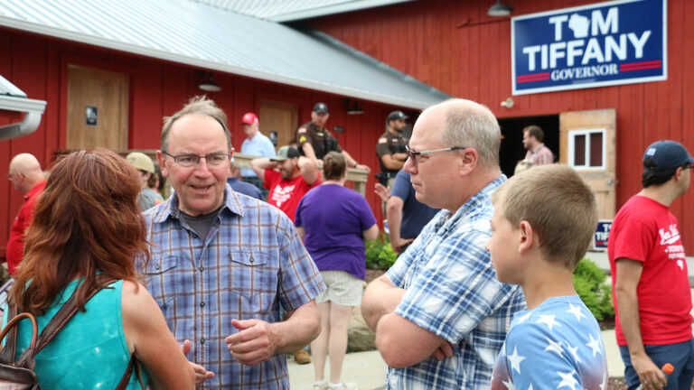 Tom Tiffany speaks to a person facing him with two other people to his side while standing outside in front of other people standing and walking in front of a building with wood siding, multiple levels of corrugated metal roofs and a sign reading Tom Tiffany Governor.